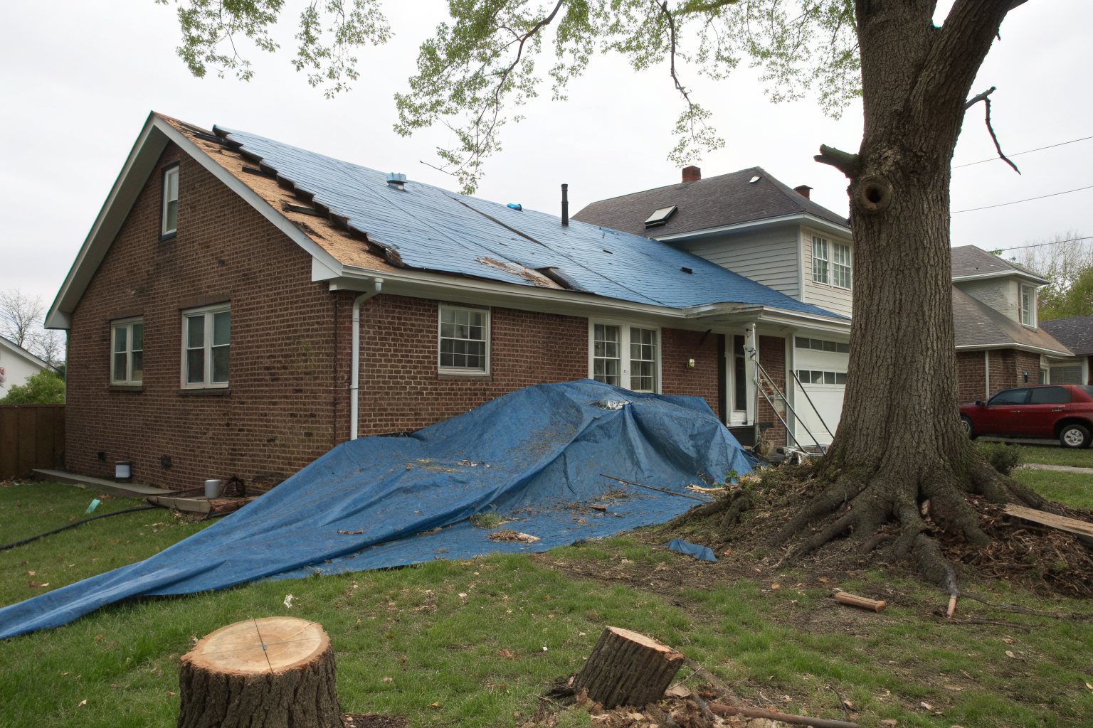 A blue tarp over part of a brick house roof beside a cut tree stump